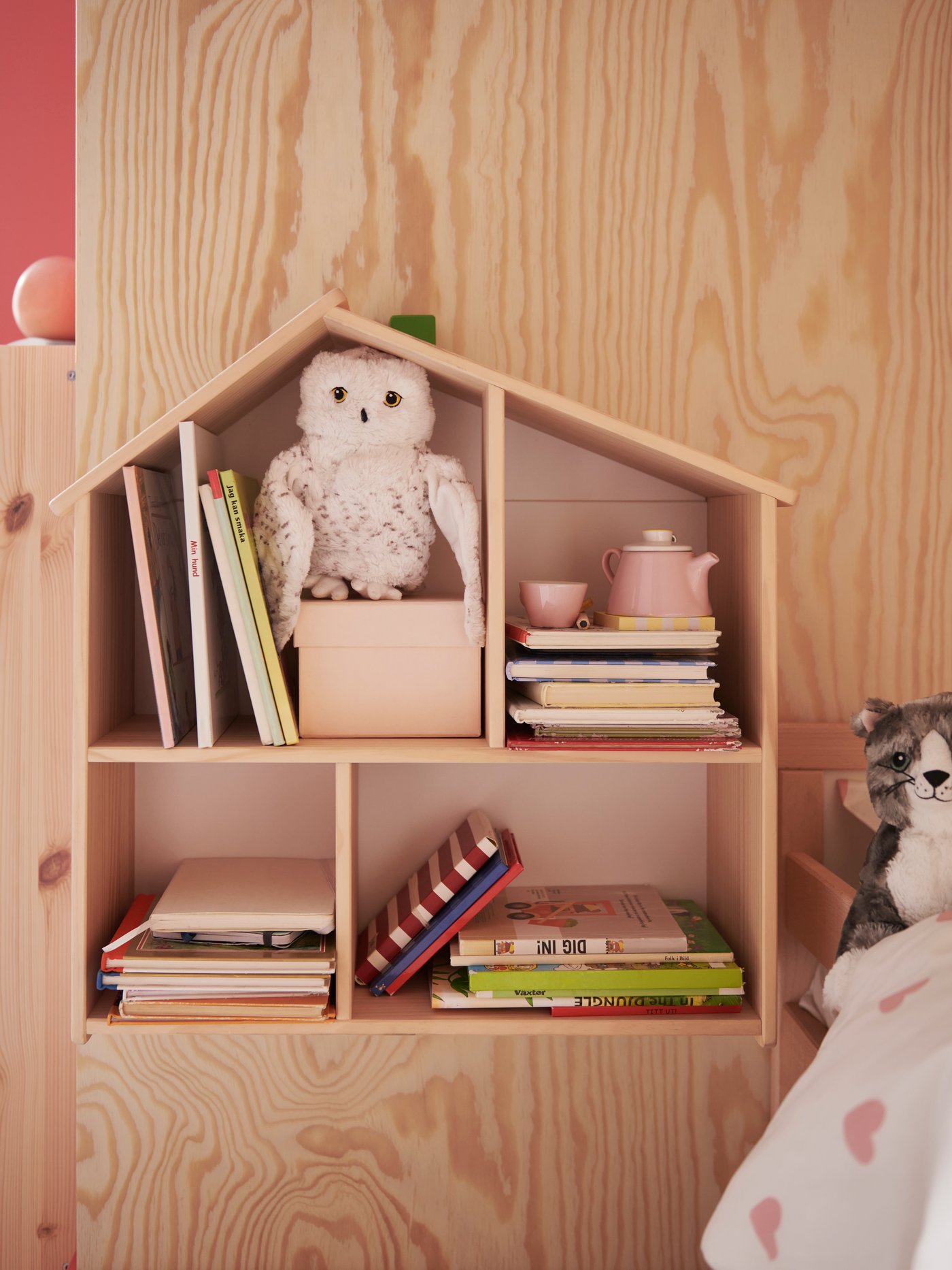 A FLISAT doll’s house/wall shelf on a wooden wall by a bed holds books, a box and a snowy owl SKOGSDUVA soft toy hand puppet.