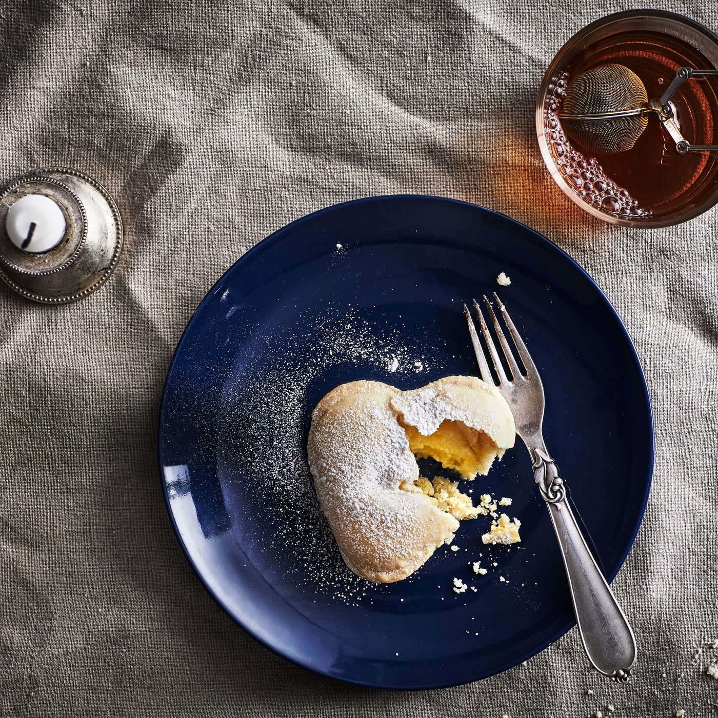 A vanilla heart biscuit and a fork on a dark blue plate, a mug with tea and a candle.