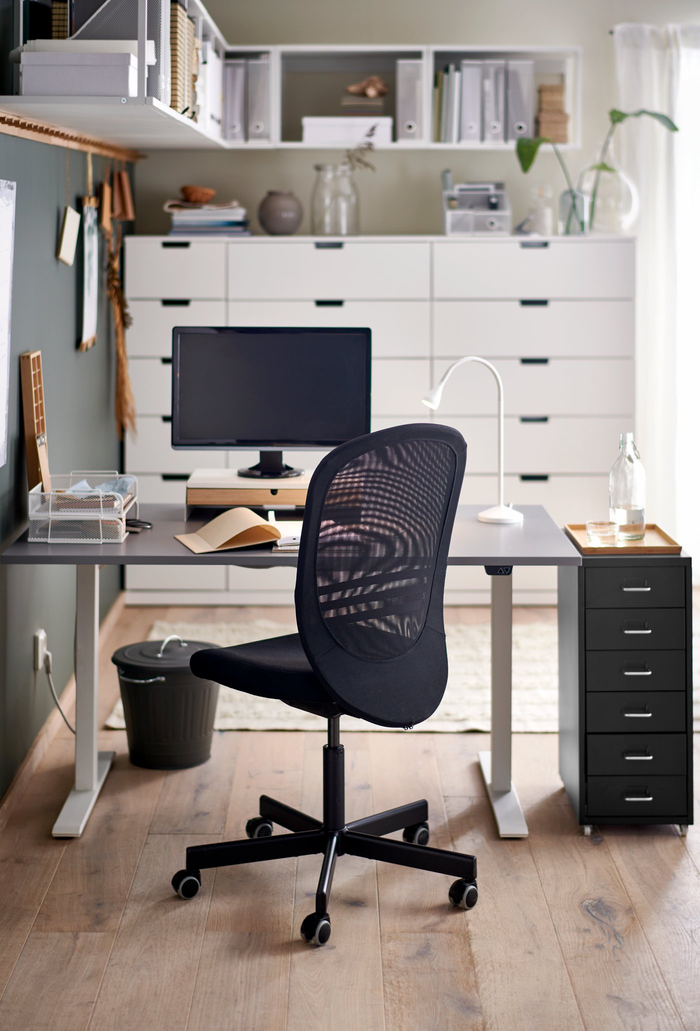 A work station with RODFUL sit/stand desk in grey/white and a black mesh office chair faces a wall of white cabnets.