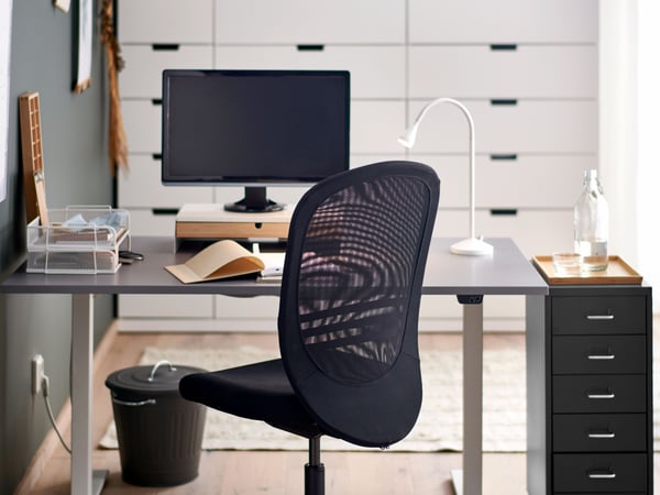 A black FLINTAN office chair and HELMER drawer unit by a grey RODULF sit/stand desk with a work lamp and computer screen.