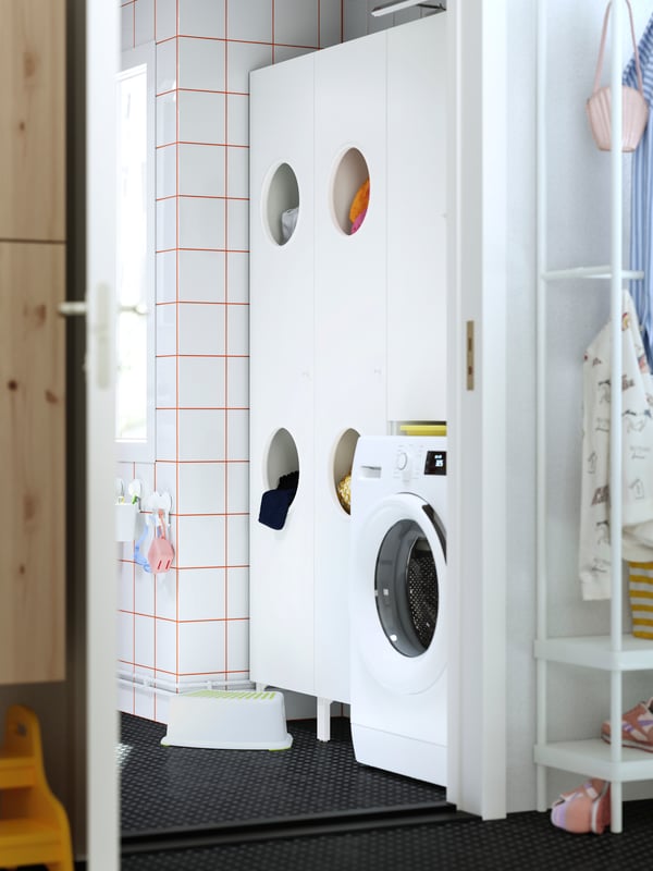 A laundry room with a NYSJÖN laundry cabinet with openings for sorting clothes, next to a washing machine and step stool.