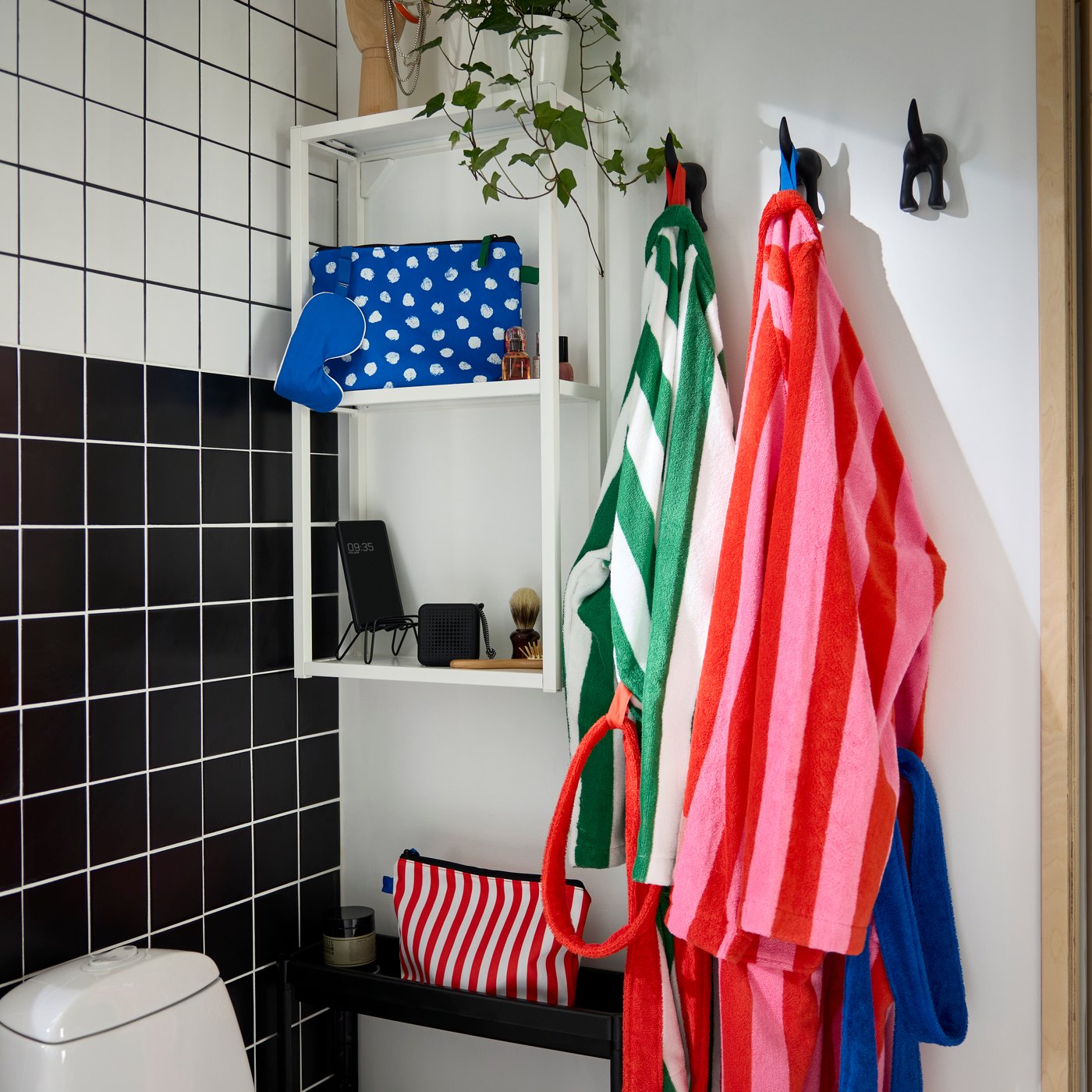 In a corner of a bathroom is a white ENHET wall shelf with a blue toiletry bag and grooming tools. Two striped bathrobes.