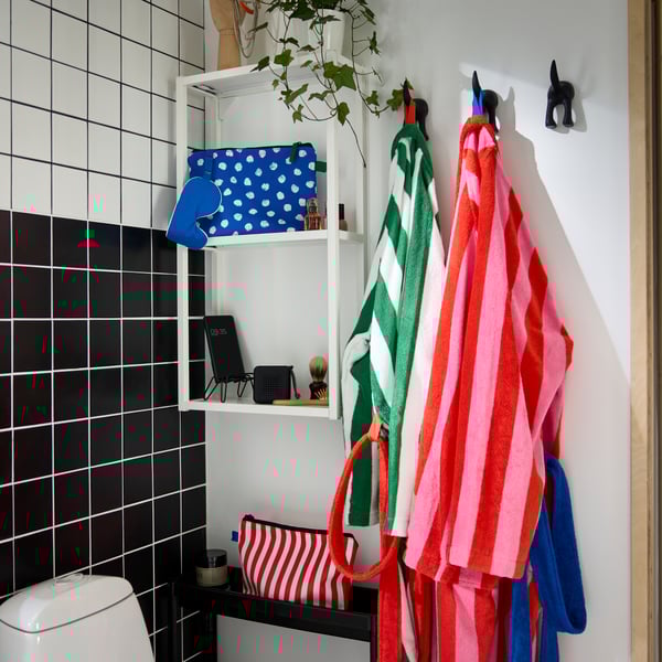 In a corner of a bathroom is a white ENHET wall shelf with a blue toiletry bag and grooming tools. Two striped bathrobes.