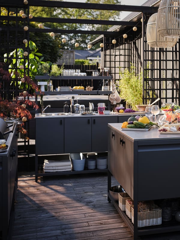 Sonnige Terrasse mit Grünpflanzen und drei SOLVINDEN LED-Hängeleuchten über einer vollständigen BÅTSKÄR Außenküche.