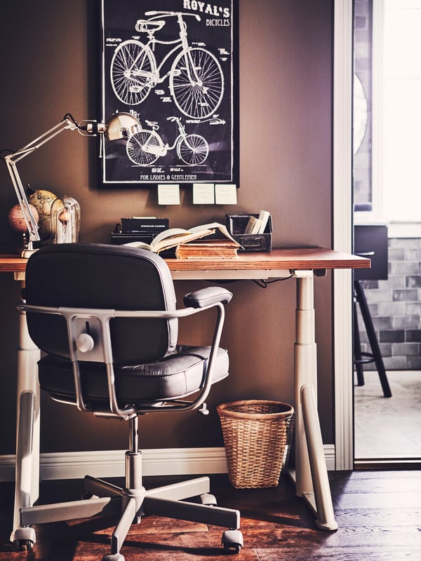 An office chair in front of a brown-beige IDÅSEN sit/stand desk with books and a nickel-plated FORSÅ work lamp.