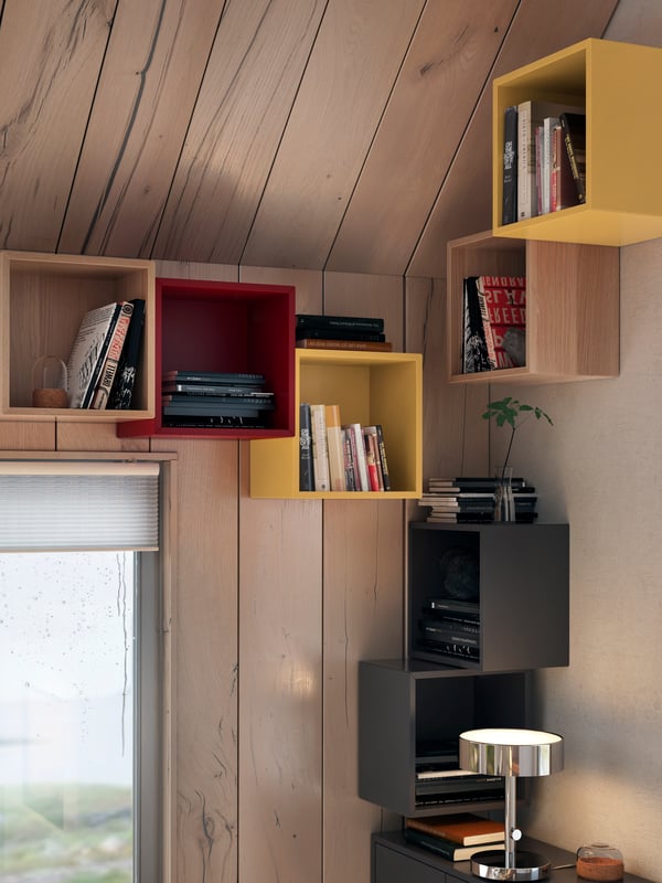 Estantes modulares EKET de colores madera, rojo, amarillo y negro con libros y adornos en la intersección de 2 paredes con el techo.