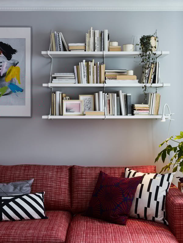 Red sofa with white and red patterned decorative cushions. White shelves are mounted on the wall behind and store books.