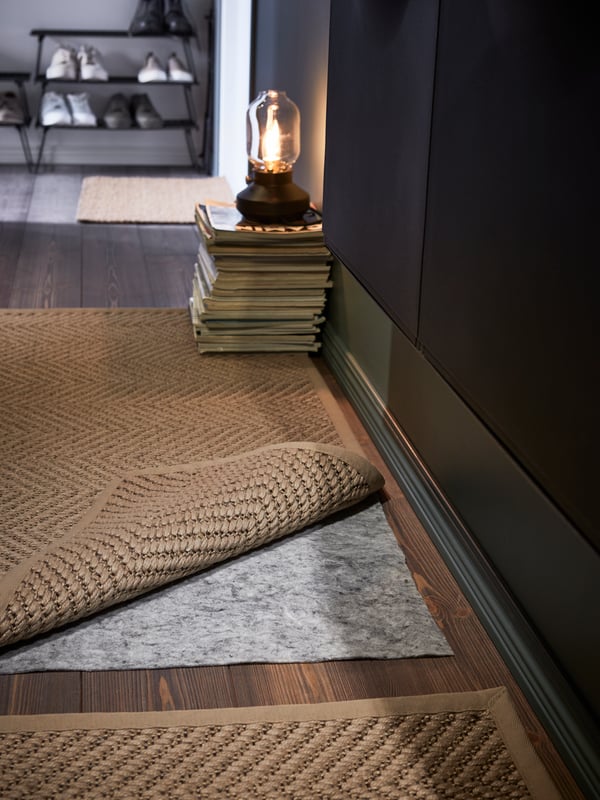 Zwei Teppiche liegen in einer Reihe in einem Flur. Die Kante eines Teppichs ist hochgeklappt und zeigt eine STOPP FILT rutschhemmende Teppichunterlage.
