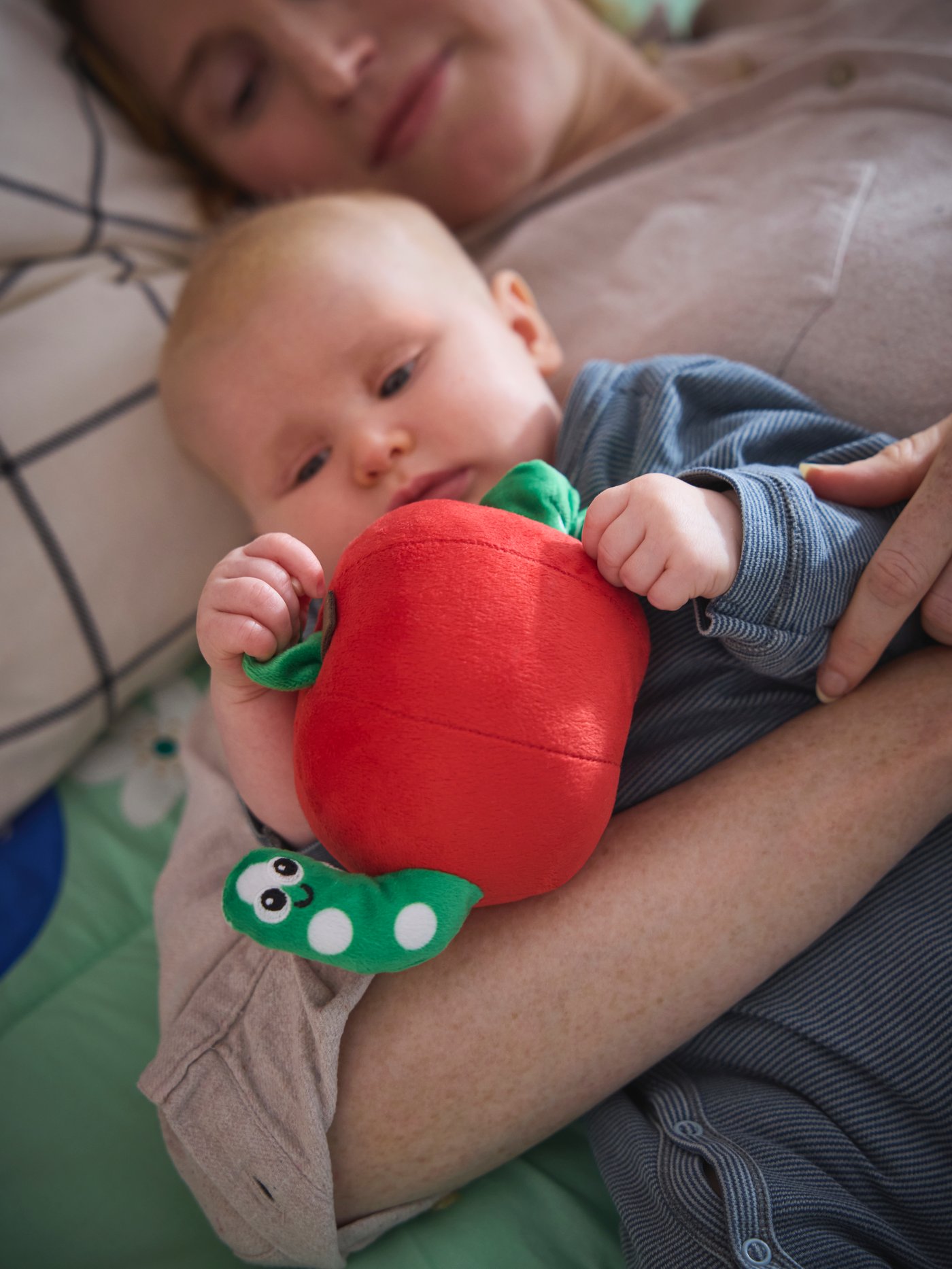 A baby laying on the floor in its mother's arms, holding a red apple SANDBI musical toy.