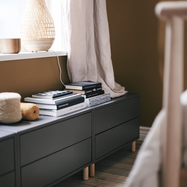 A bedroom where dark-grey EKET cabinets with two drawers stand beneath a sunlit window, books stacked on top.