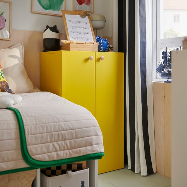 Leuchtend gelbe LASTARE Aufbewahrungskombination mit Kinderspielzeug neben einem Bett mit weiß-grüner Bettwäsche.