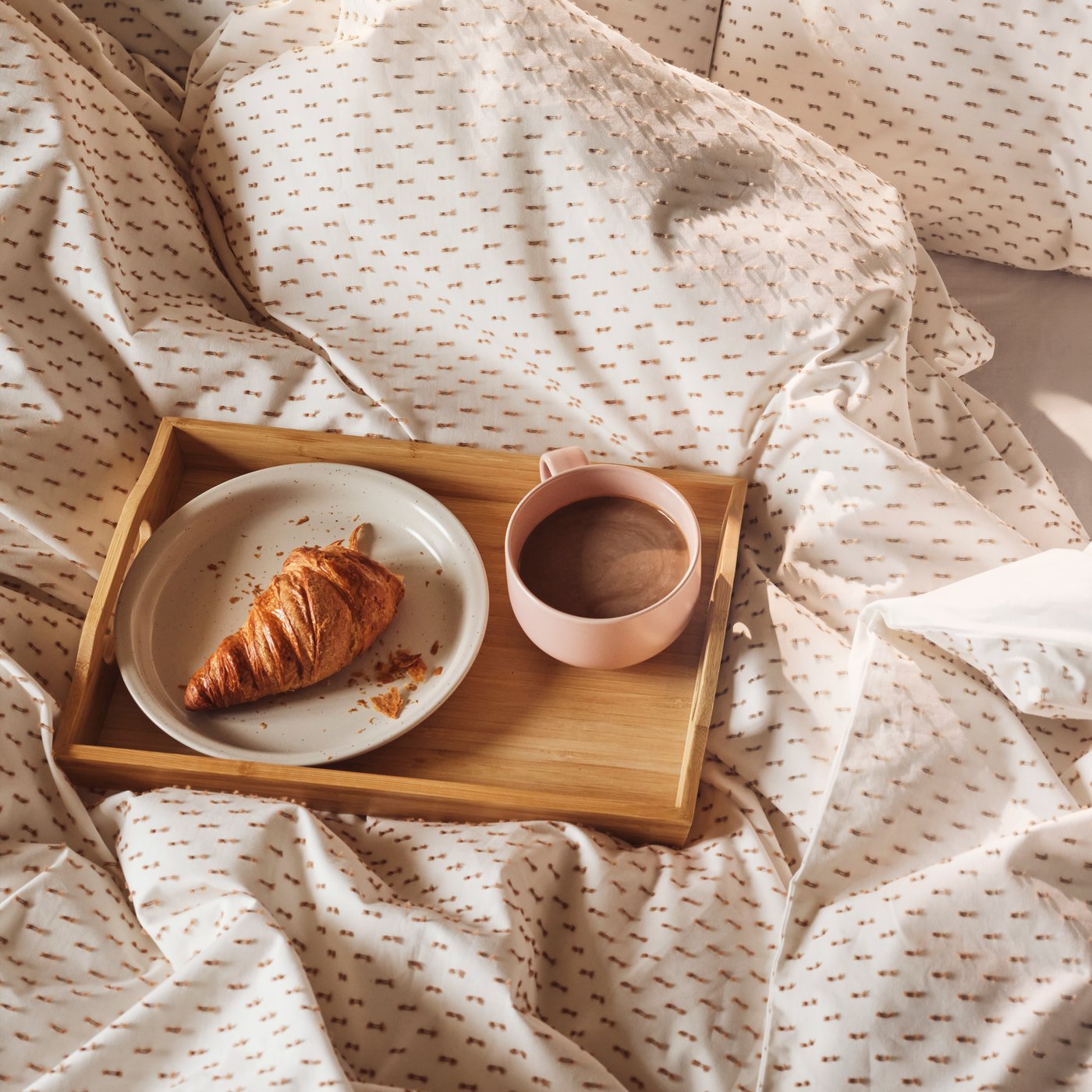 En bakke med en kaffekop og en tallerken står på en seng med lyserødt sengetøj 