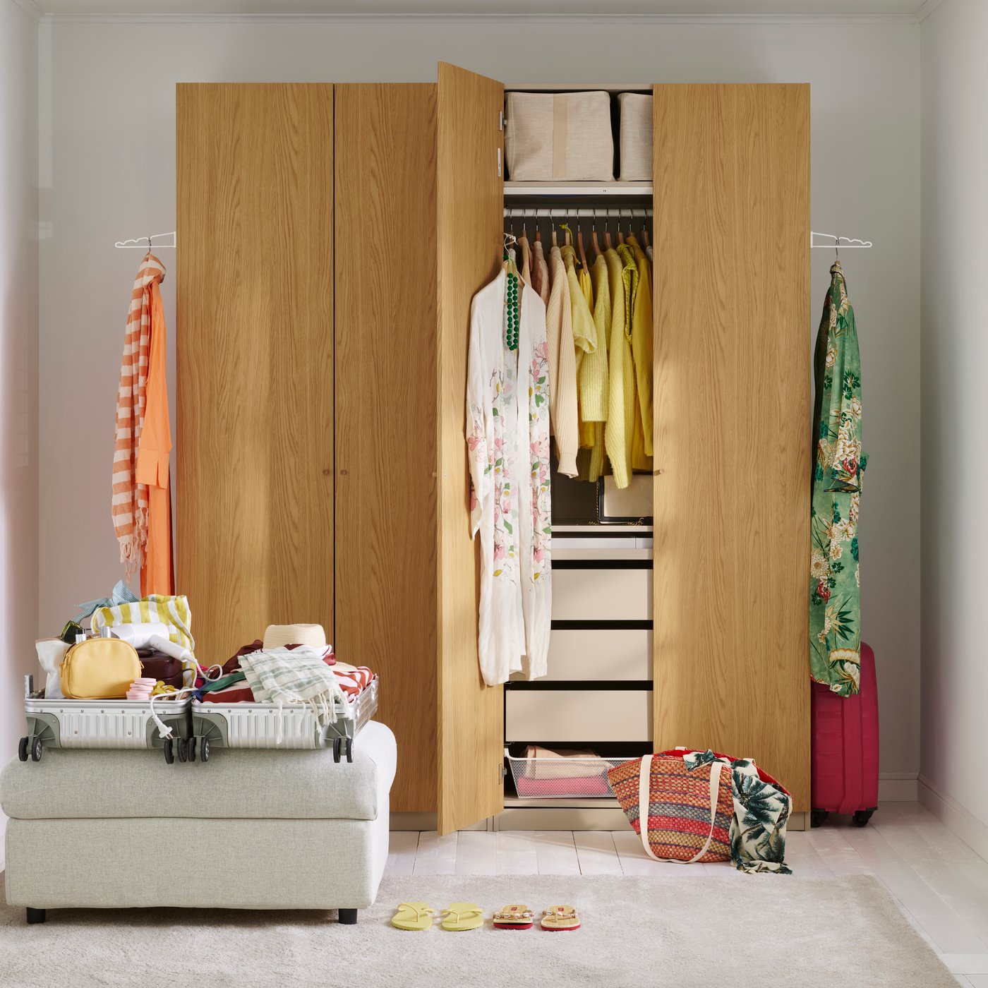 An open wooden PAX wardrobe with 3 KOMPLEMENT valet hangers attached with clothes hanging. In front is an open, half-packed suitcase.