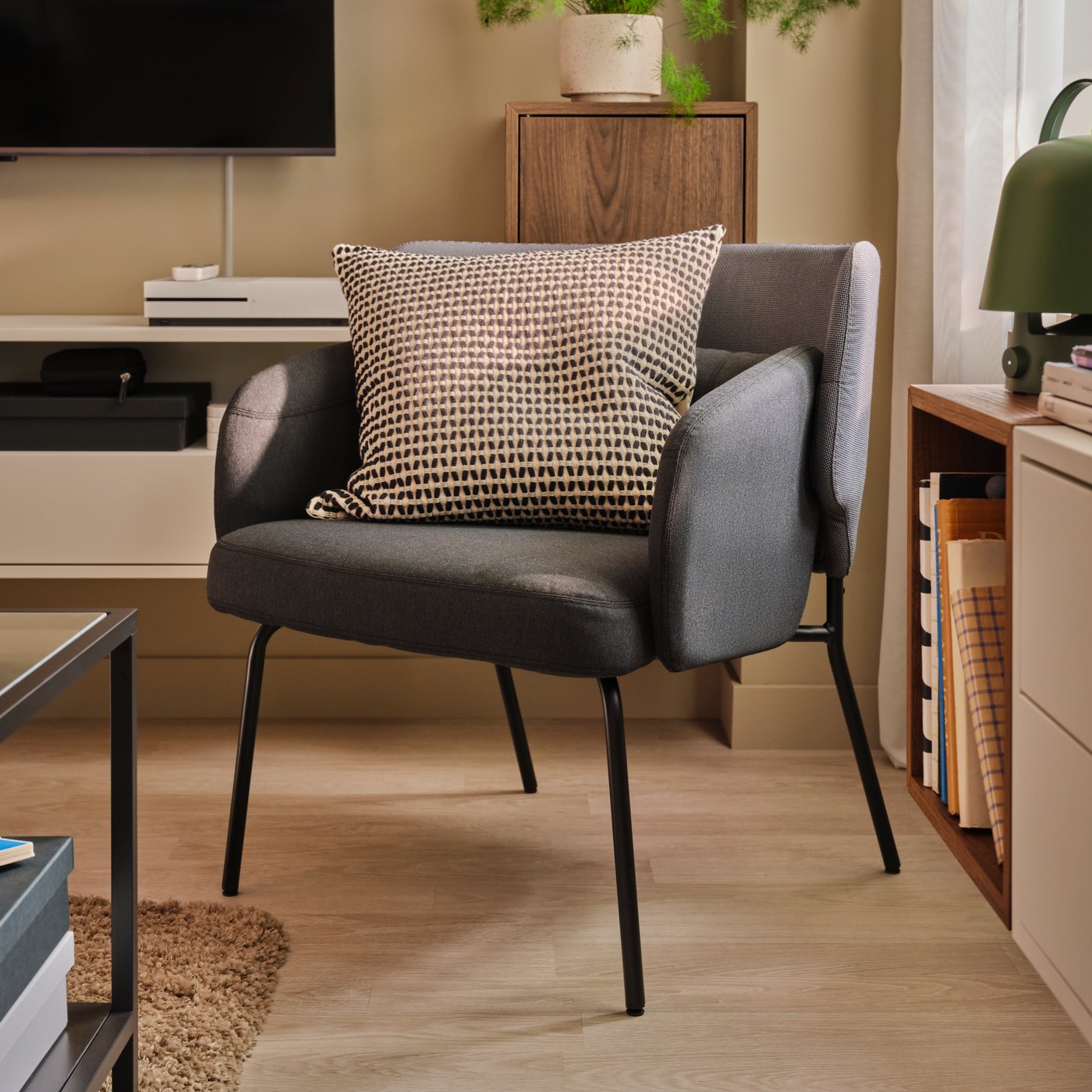 A light living room with a dark-grey BINGSTA armchair holding a cushion and placed by a TV set and wall-mounted cabinets.