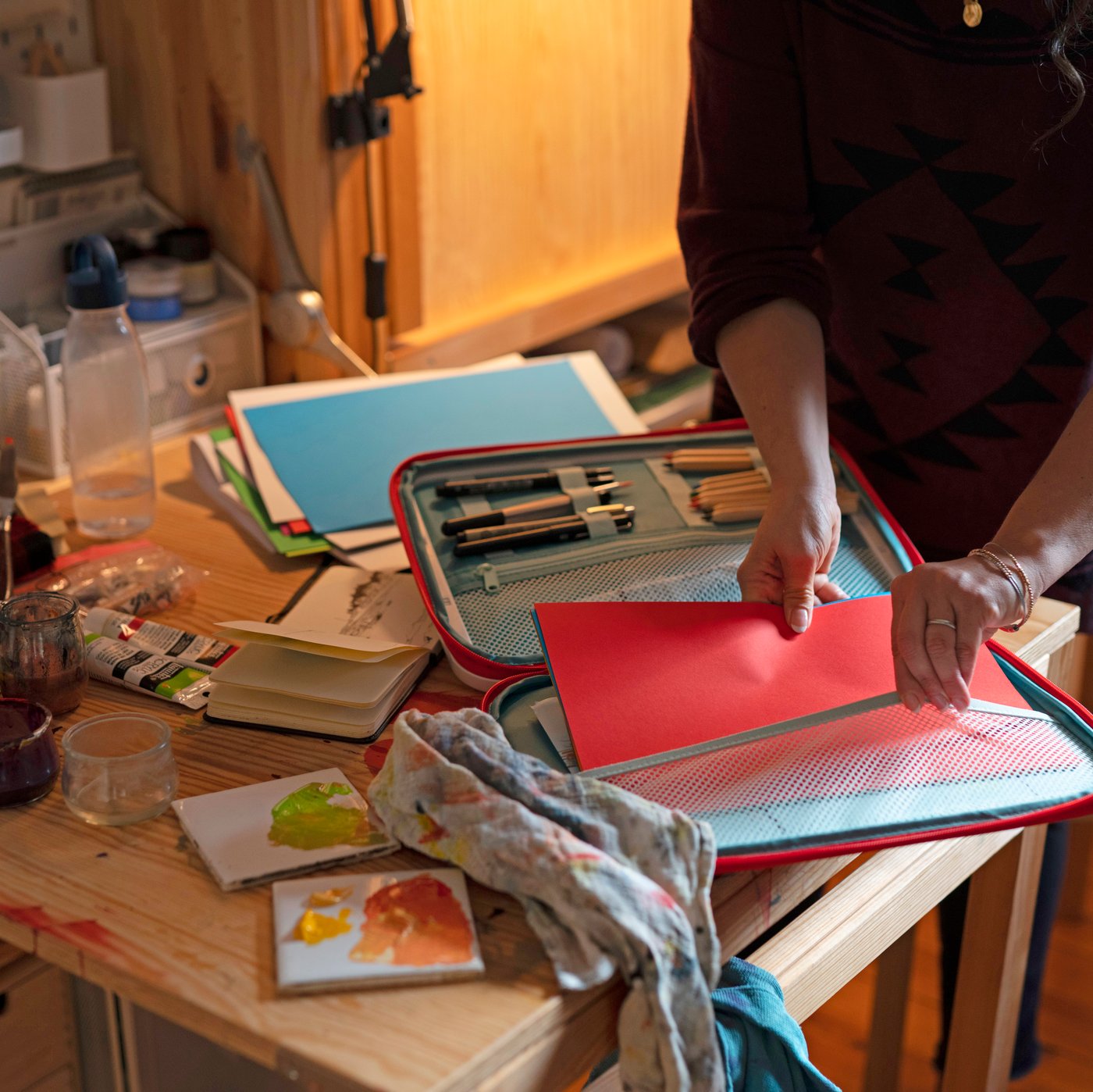 A woman in an art studio is preparing a MÅLA portable drawing case with pencils and paper.