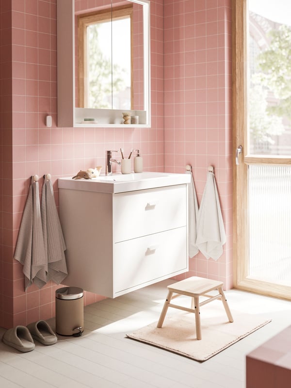 A white HAVBÄCK wash-stand with drawers and a mirror cabinet are placed in a bathroom with soft pink walls and a grey floor.