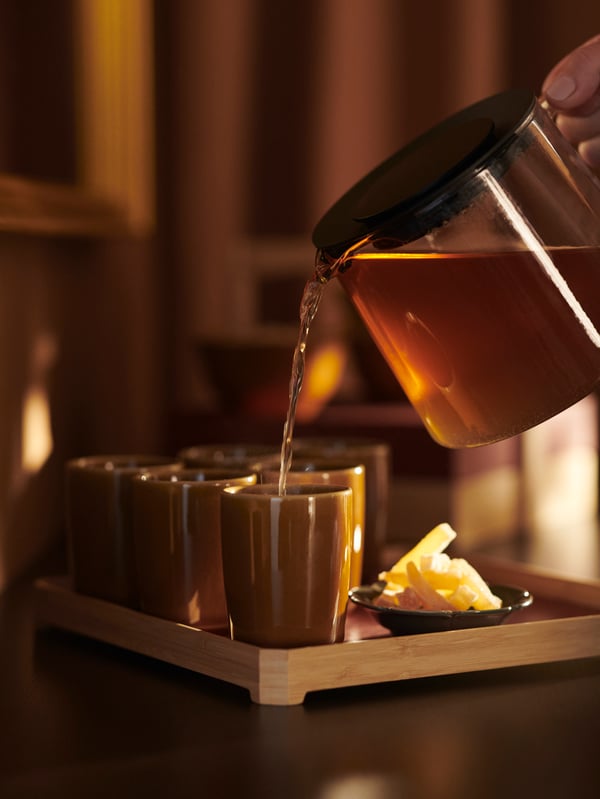 A person pours tea into yellow FÖSSTA mugs placed on a bamboo tray.