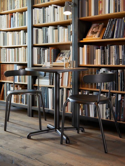 O masă rotundă cu două scaune gri este așezată în fața mai multor biblioteci cu cărți pe rafturi.