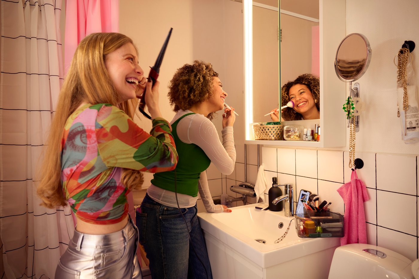 Two women in party outfits are putting on makeup and curling their hair in a small bathroom.