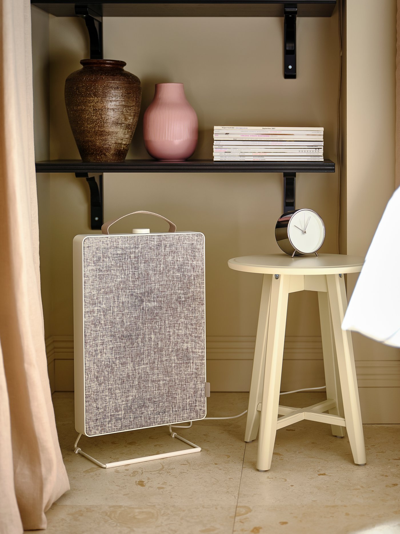 Luftrenare med tygfront står bredvid ett litet träbord med klocka, under en hylla med keramikvaser och böcker i ett ljust rum.