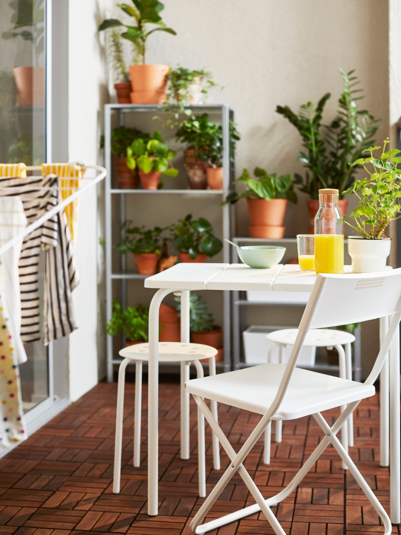Una mesa VÄDDÖ blanca, frente a una silla plegable FEJAN y dos pisos MARIUS. La terraza tiene pisos deck de madera RUNNEN y al fondo un estante HYLLIS con plantas.