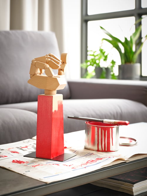 A wooden LÅNESPELARE accessories stand on a table in front of a grey sofa, in the process of being painted red.