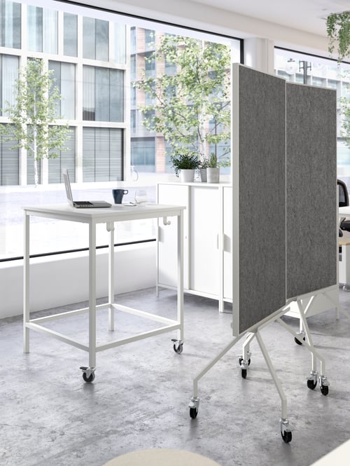 A modern workspace that features a white underframe with castors and a table top next to a noticeboard on castors. 