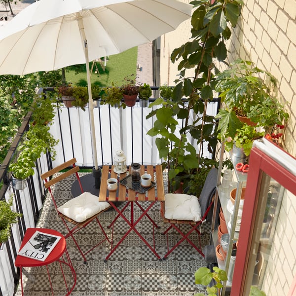 Un pequeño balcón con muchas hierbas y plantas, una estantería con macetas, un parasol, dos sillas y una mesa.
