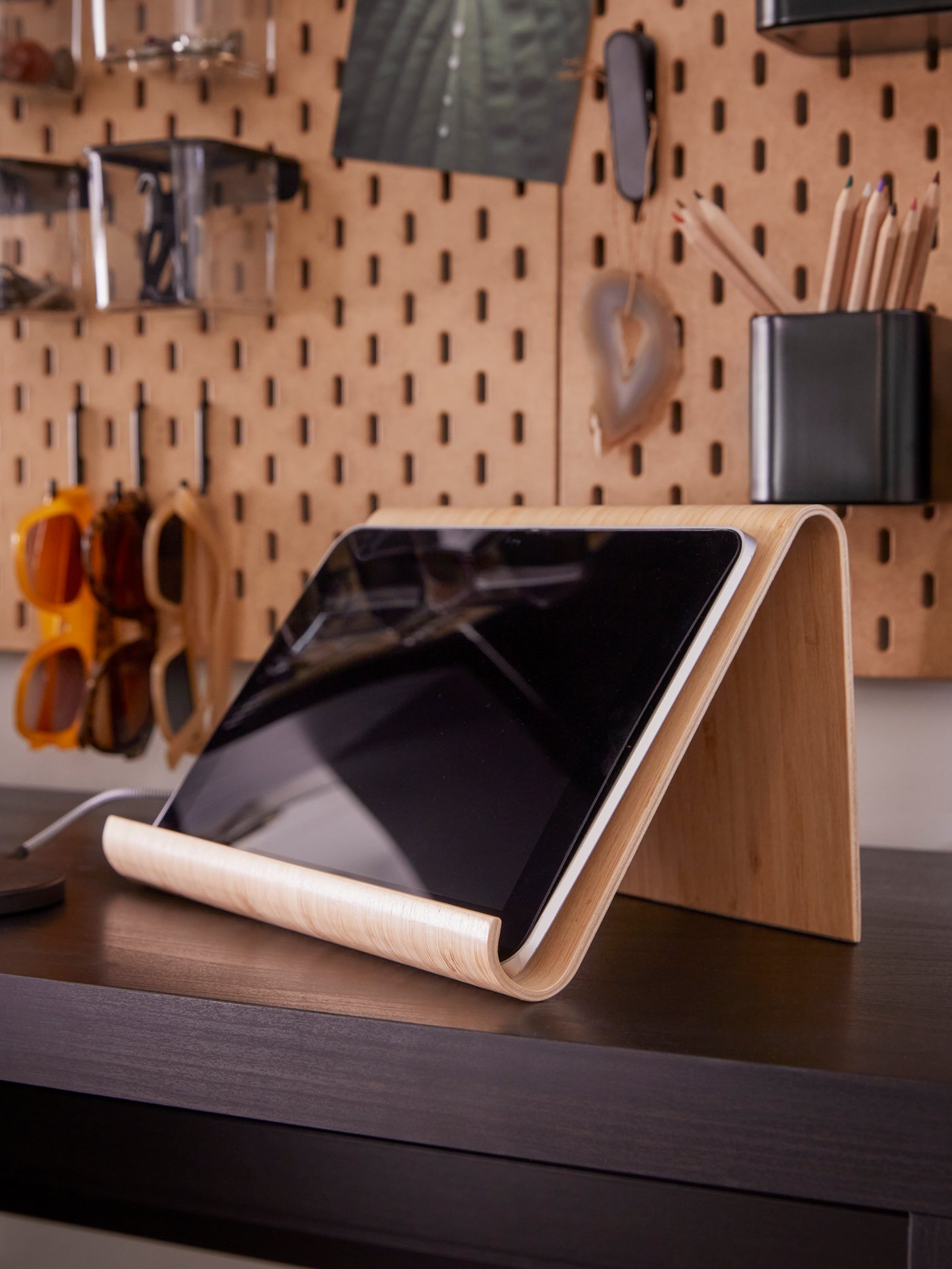 A VIVALLA bamboo veneer tablet stand with a tablet on it is on a desk, with a pegboard with accessories hanging above it.