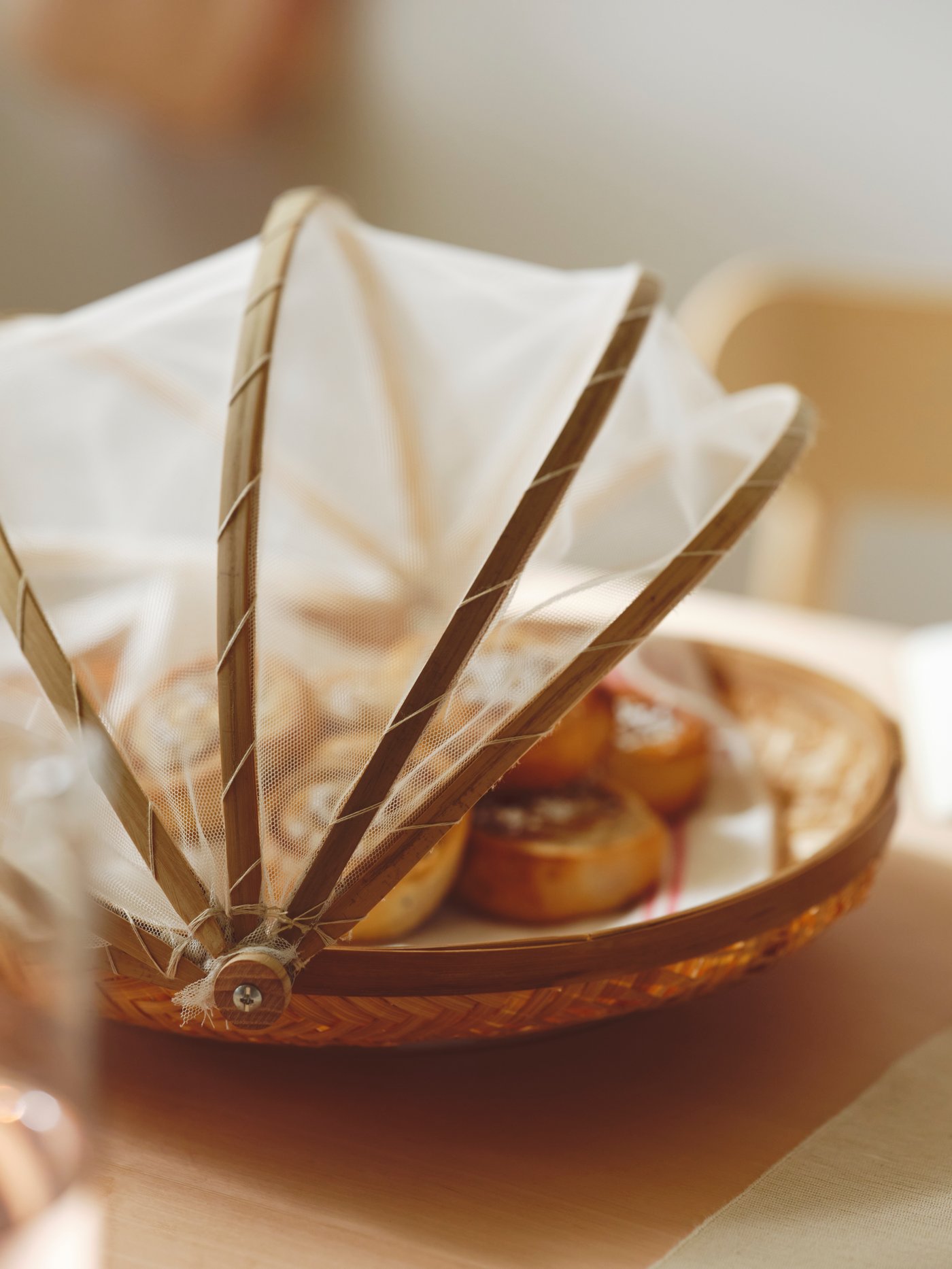 A table with a SNUSTORR food cover with tray in handmade bamboo holding several KAFFEREP cinnamon buns.