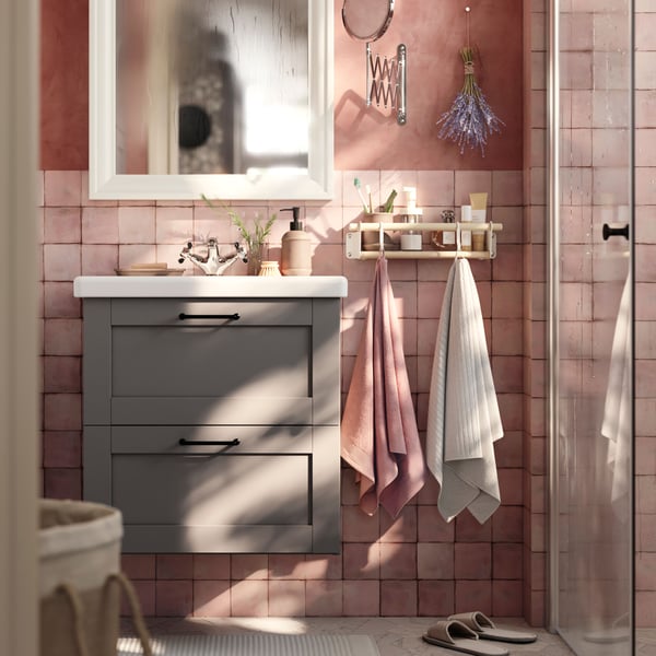 A grey HAVBÄCK wash-stand with drawers and a mirror cabinet are placed in a bathroom with soft pink walls and a grey floor.