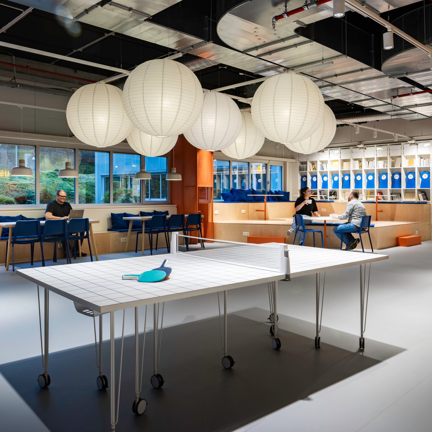 Espace de détente ouvert au bureau avec une table de ping-pong, des suspensions sphériques blanches, des chaises bleues et un mur d’étagères, où des collaborateurs font une pause.