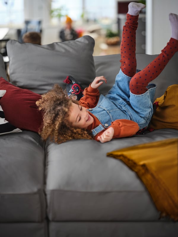 A small child in a denim dress with red leggings is laying down on a dark grey couch.