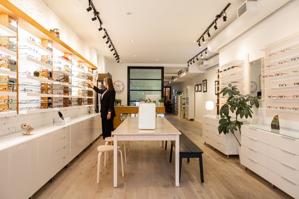 Ljus optikerbutik med långa hyllor fyllda med glasögon, ett centralt bord och en person som justerar en produkt på väggen.
