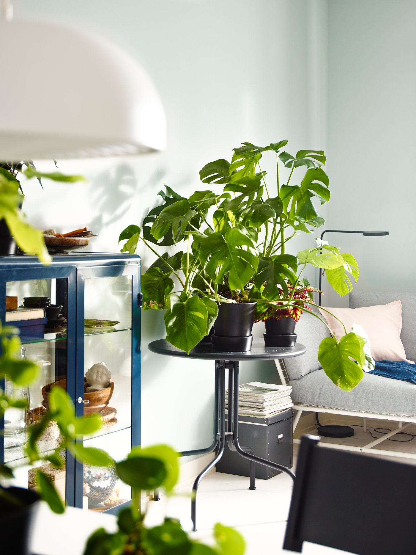 Una mesa de exterior circular y gris, LÄCKÖ, sostiene plantas en macetas junto a un sillón bajo y un armario con puertas de cristal en una zona interior.
