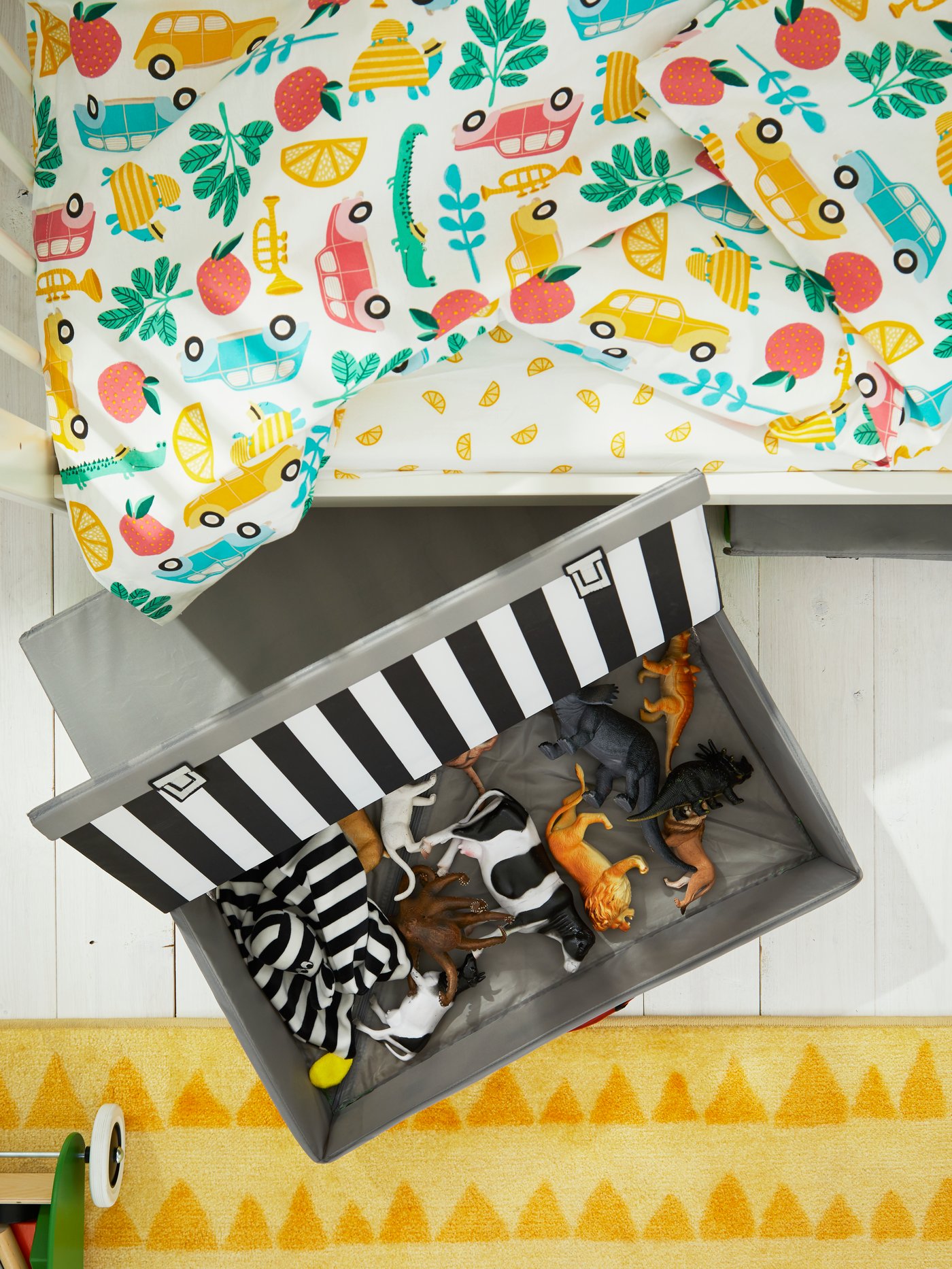 A FLYTTBAR trunk for toys containing toy animals lies on the floor beside a GULLIVER cot with RÖRANDE bed linen.
