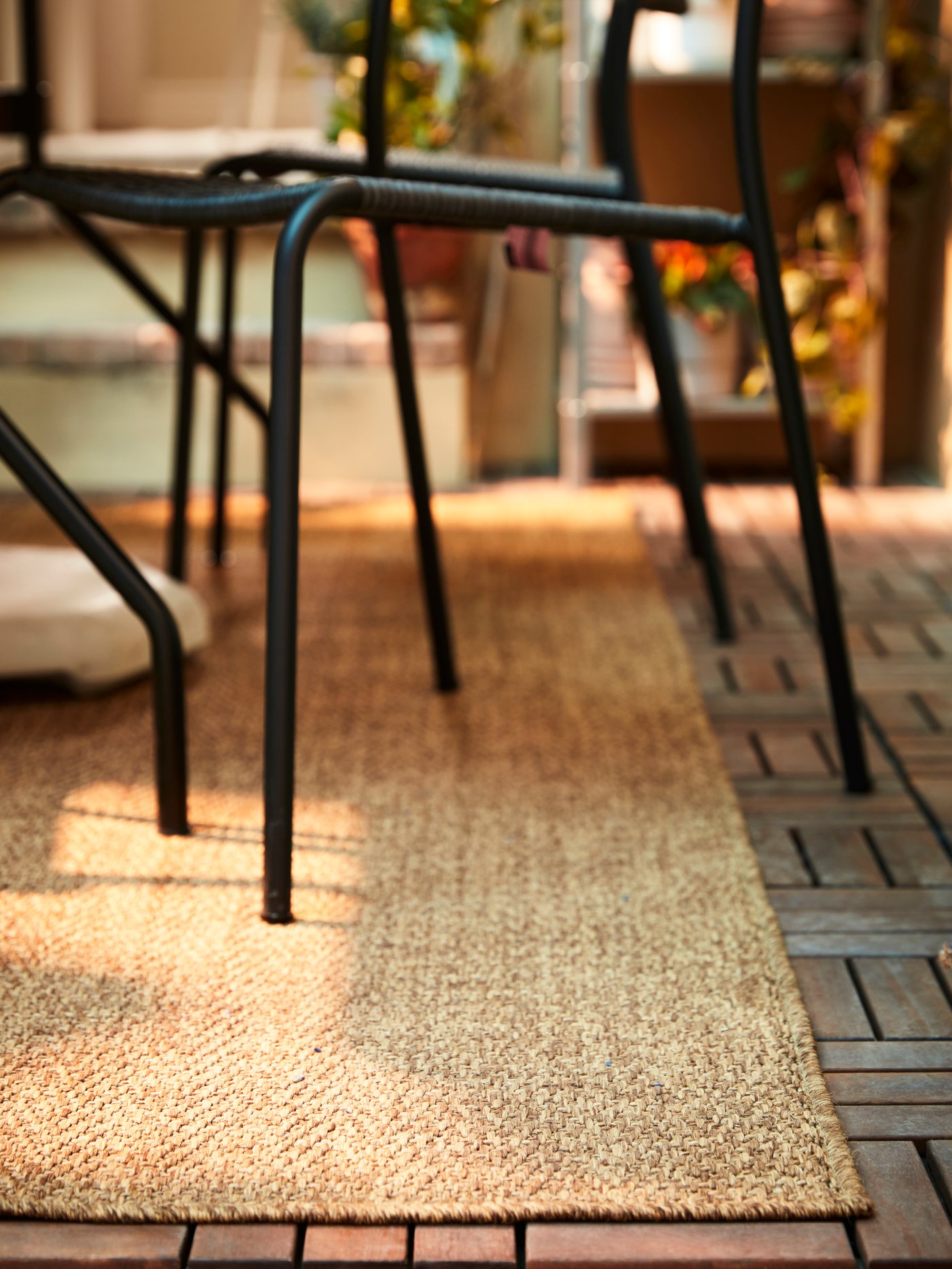 Vista de cerca de una alfombra LYDERSHOLM de aspecto rústico para uso exterior, sobre un piso deck de madera RUNNEN. Sobre la alfombra se ve una silla metálica VIHOLMEN negra.