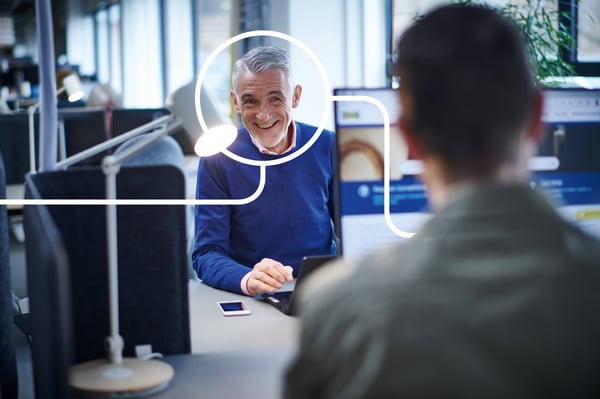 A man wearing a blue sweater is sitting across from another man who is in front of a computer screen.