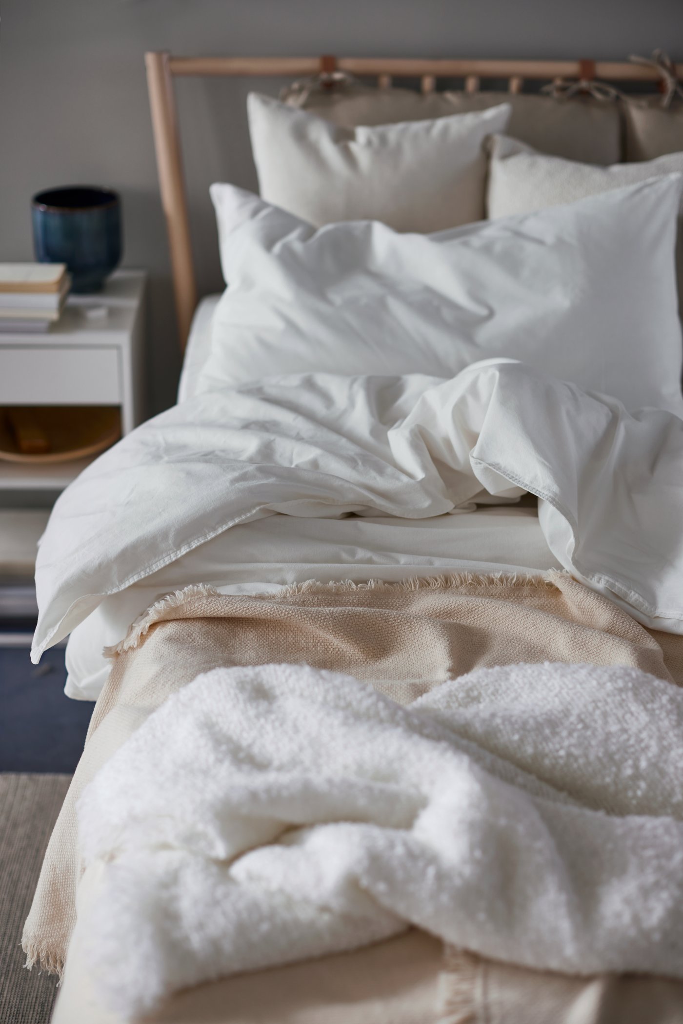 En seng med et sengestel af birk, hvidt ÄNGSLILJA sengetøj og en beige plaid står ved siden af et hvidt sengebord, hvor der ligger en stak bøger.