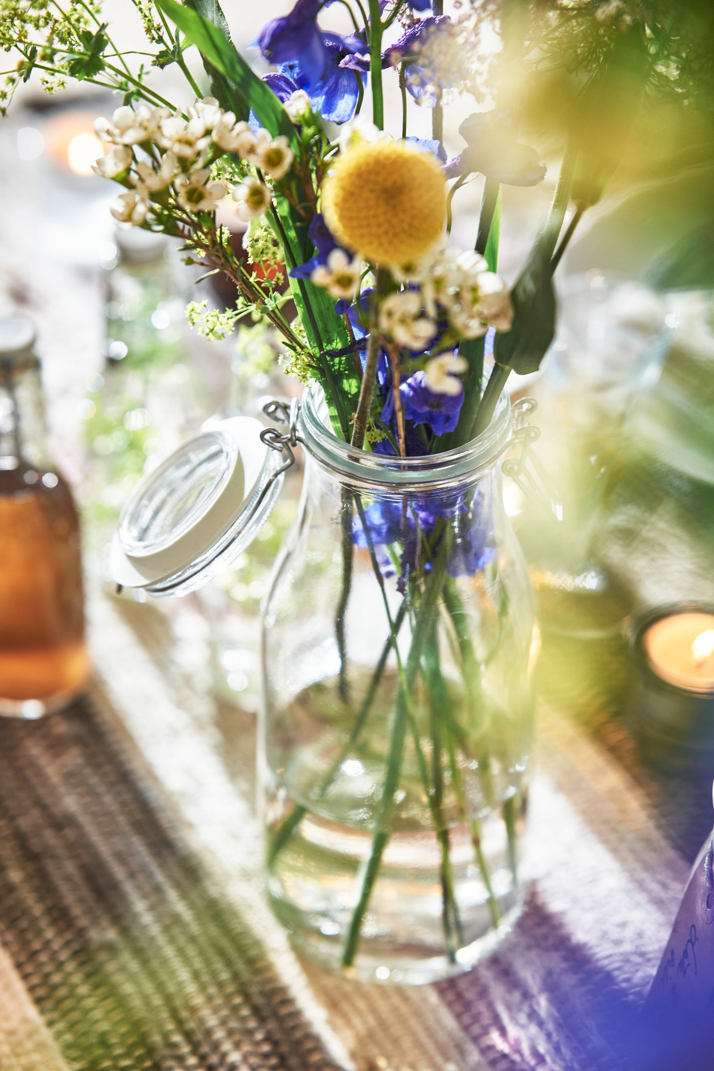 Glasflasche mit bunten Blumen auf einen Tischläufer, Kerzenlicht im Hintergrund.