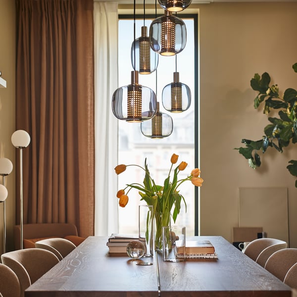 A large MÖRBYLÅNGA table and some chairs stand under two HÖGVIND pendant lamps with 3 lamps in a dining room.