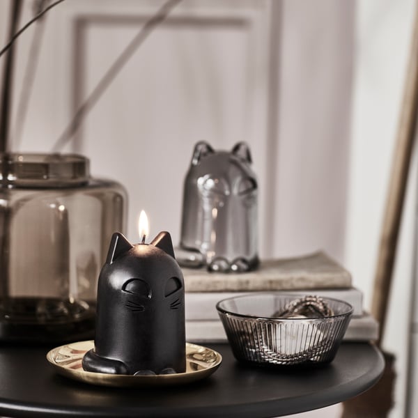 A KUSTFYR unscented candle and a KUSTFYR glass dome, both shaped like cats, are placed on a black table next to a bowl.