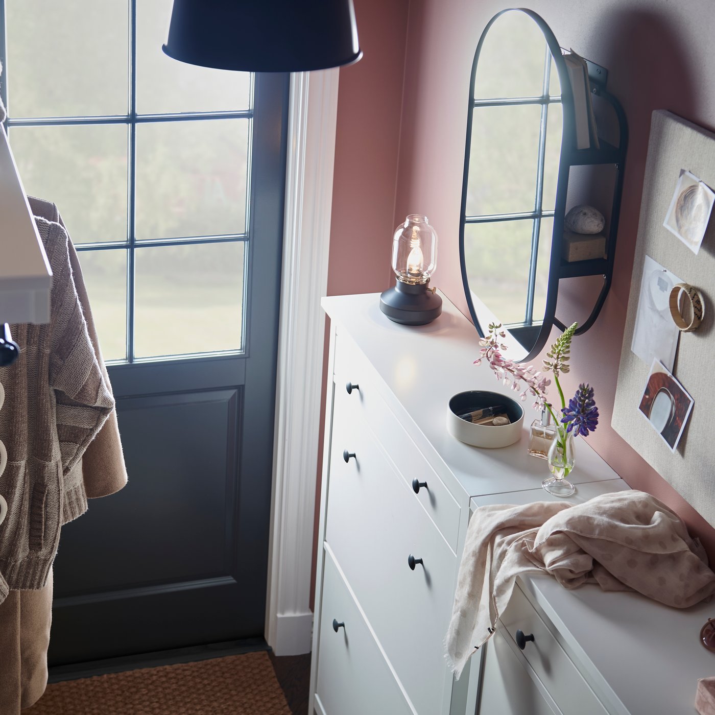 Smalt hallutrymme med rosa väggar, vit förvaring, rund spegel och krokar för kläder, skapar en organiserad och välkomnande entré.