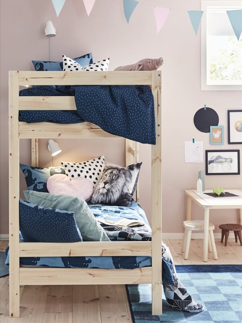 Cadre de lit superposé en pin massif MYDAL, avec beaucoup de linge de lit et de coussins bleus sur chaque lit, près d’une petite table dans une chambre d’enfants.