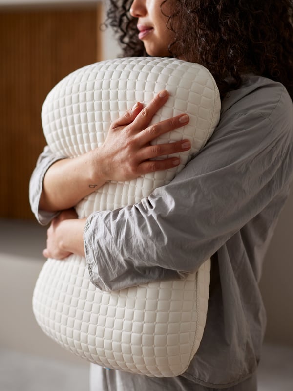 A woman in grey pyjamas hugging a KLUBBSPORRE ergonomic pillow.