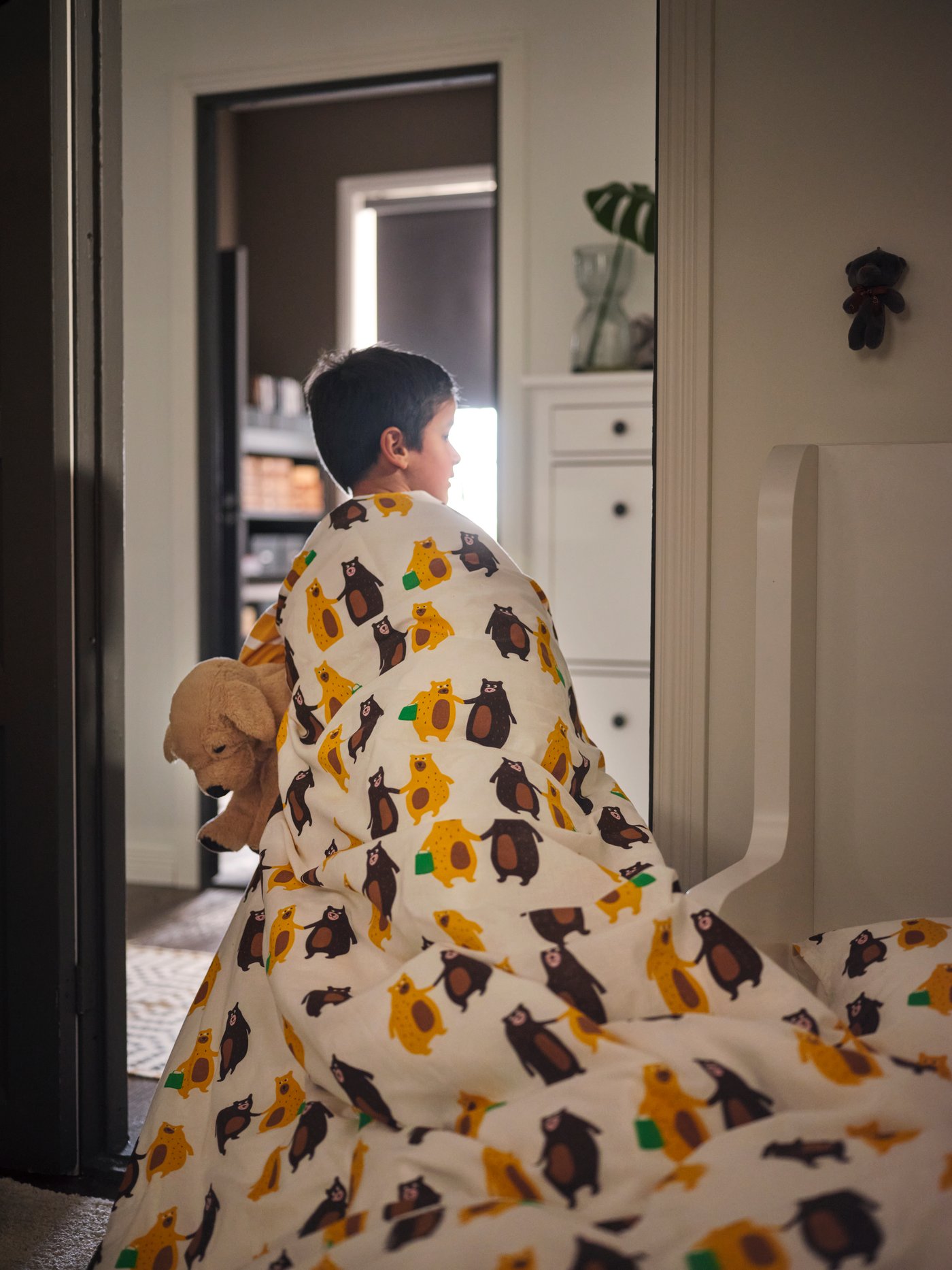 طفل يمشي على سجادة حياكة مسطحة VAMDRUP باتجاه باب غرفة نوم ممسكًا بدمية دب وملفوفًا ببياضات السرير BRUMMIG.