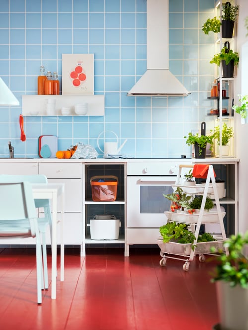 A light blue tiled wall with a white ENHET kitchen, ENHET shelves, a RISATORP trolley, a kitchen table, herbs and conserves.