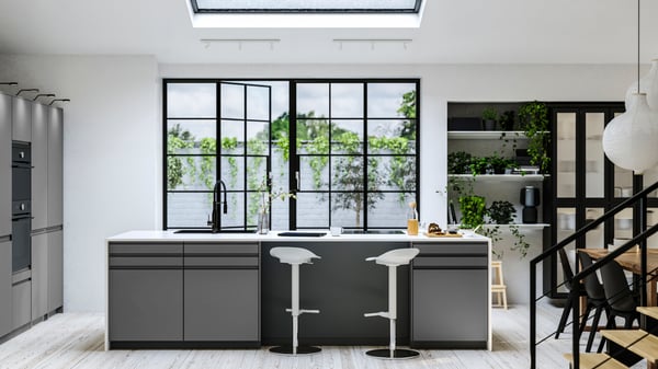 Ett modern kök med ett stort fönster som vetter mot grönska. Köket har en central köksö med två barstolar, eleganta grå skåp och vita bänkskivor. Det finns växter på hyllorna till höger och en matplats delvis synlig längst till höger. Utrymmet är väl upplyst av naturligt ljus som kommer in genom takfönstret ovanför.