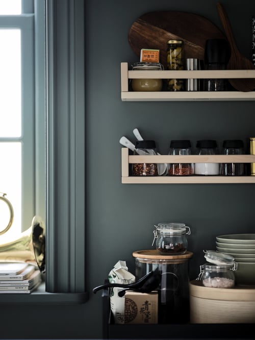 Une paire de supports à épices BEKVÄM en tremble remplis de condiments et montés sur un mur de cuisine vert à côté d’une fenêtre.