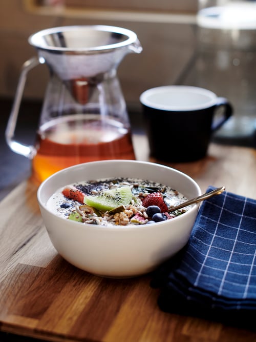 Wooden tray with white porcelain bowl with cereal and fruit, glass teapot with strainer, black mug and blue cloth napkin.